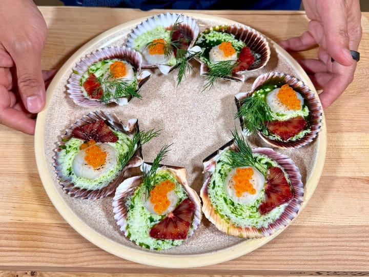 Scallops in half shells topped with roe, dill, and blood orange slices, served with green herb foam on a ceramic plate, held by hands over a wooden surface.